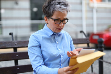 A stylish woman carefully reads a letter or notice from an envelope while sitting on a street bench.