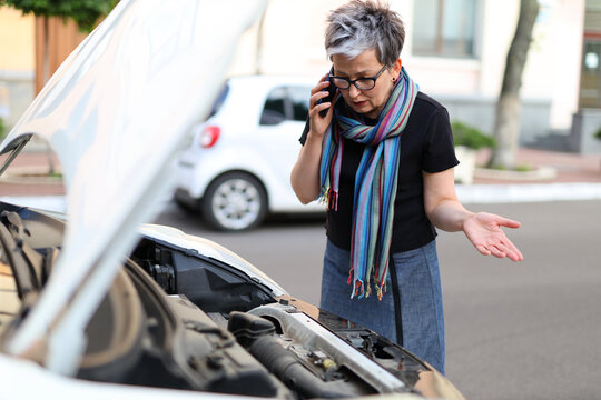 A woman stands in front of a car with an open hood and call to service department or calls a tow truck.