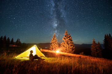 Night camping in mountains under starry sky. Silhouette of tourist sitting near tent in campsite,...