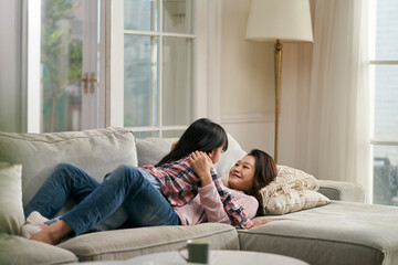 Obraz premium happy asian mother and daughter lying on couch and chatting at home