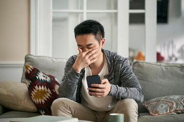 young asian man sitting on couch at home feeling eye fatigue as result of cellphone use