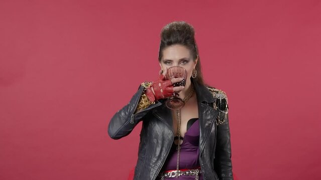 A woman holds a glass of wine, takes a sip, feeling its aftertaste with pleasure exhales. A woman drinks wine, raises her glass in toast. A woman on a red background. HDR BT2020 HLG Material.