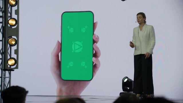 A woman presenter is standing on a well-lit stage in front of a large LED screen showing a hand holding a generic smartphone with green screen. Technology, application or product launch presentation 