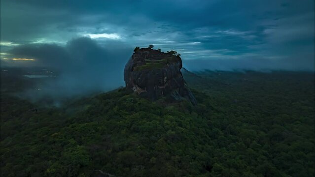 Aerial footage of ancient rock fortress towering above surrounding forest. Hyperlapse shot of clouds rolling above landscape at dusk. Sigiriya, Sri Lanka