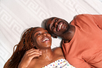 An elated African couple, a man and a woman, lie on a bed in a top-down shot, exuding happiness and...