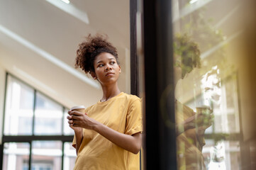 Young African American woman relaxing In modern office looking out of Window with hot drink.