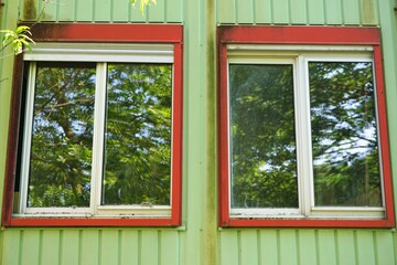 Containerfassade mit grüner Wand aus Metall und zwei Fenstern mit weißen Holzrahmen und roten Metallrahmen bei sonne am Mittag im Sommer