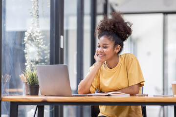 Business And Education Concept. Smiling african american  sitting at desk working on laptop writing letter in paper documents, free copy space. Happy millennial female studying using laptop	