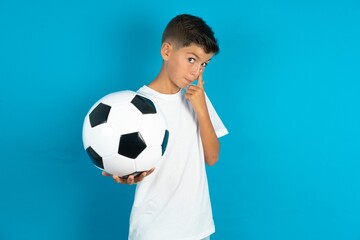 Little hispanic boy wearing white T-shirt holding a football ball Pointing to the eye watching you gesture, suspicious expression.