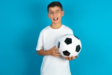 Little hispanic boy wearing white T-shirt holding a football ball sticking tongue out happy with funny expression. Emotion concept.