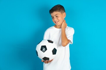 Little hispanic boy wearing white T-shirt holding a football ball Suffering of neck ache injury, touching neck with hand, muscular pain