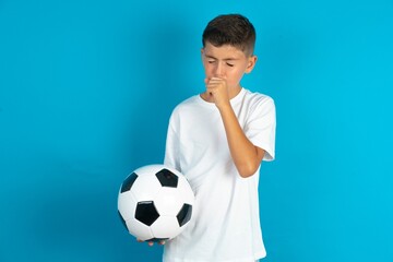 Little hispanic boy wearing white T-shirt holding a football ball feeling unwell and coughing as...