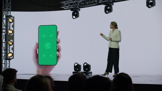 A woman presenter is standing on a well-lit stage in front of a large LED screen showing a hand holding a generic smartphone with green screen. Technology, application or product launch presentation 