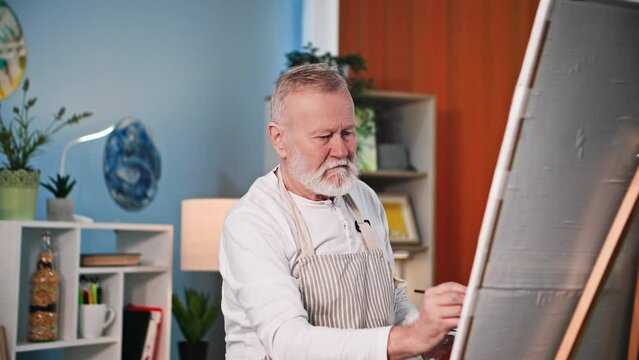elderly people hobby, creative old man draws a picture with paints and brushes using an easel in room