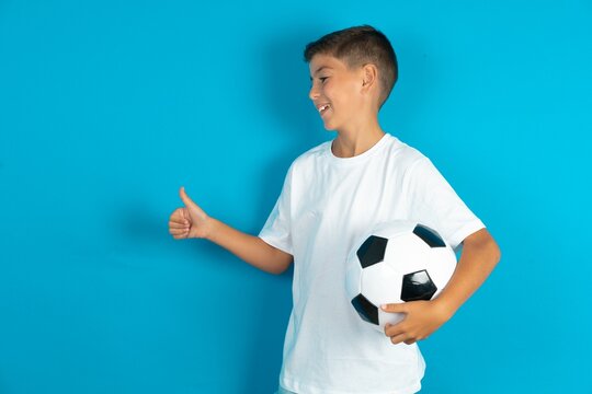 Little hispanic boy wearing white T-shirt holding a football ball Looking proud, smiling doing thumbs up gesture to the side. Good job! - Powered by Adobe