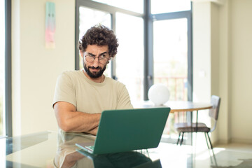 young adult bearded man with a laptop feeling displeased and disappointed, looking serious, annoyed and angry with crossed arms