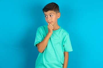 Thoughtful Little hispanic boy wearing green T-shirt holds chin and looks away pensively makes up great plan