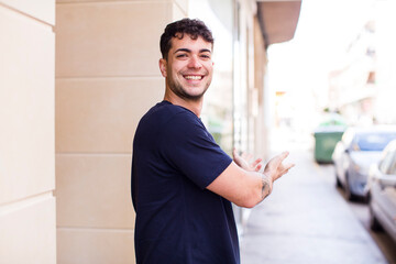 young handsome man feeling happy and cheerful, smiling and welcoming you, inviting you in with a friendly gesture