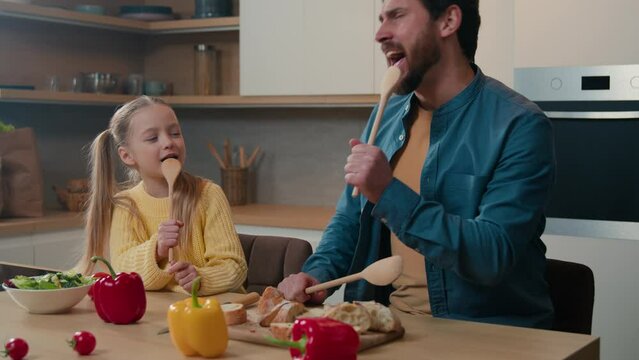 Caucasian father dad little child girl cute kid daughter happy family at home domestic kitchen singing in wooden spoons together sing song having fun fooling around playing party breakfast cooking