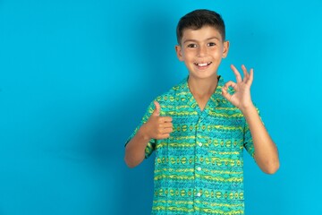 Little hispanic boy wearing green aztec shirt smiling and looking happy, carefree and positive, gesturing victory or peace with one hand