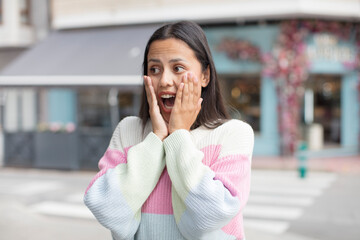 pretty hispanic woman feeling happy, excited and surprised, looking to the side with both hands on face