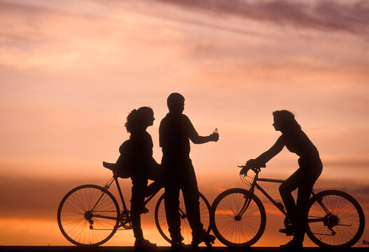 Two teenage girls in bicycles and one teenage boy with rolling skates at sunset