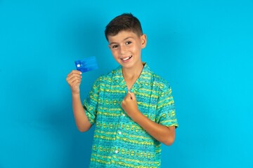 Excited happy positive cheerful smiling Little hispanic boy wearing green aztec shirt hold credit card raise fist in victory