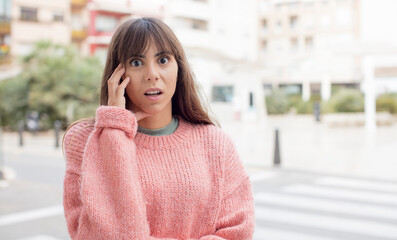 Fototapeta premium pretty young woman open-mouthed in shock and disbelief, with hand on cheek and arm crossed, feeling stupefied and amazed