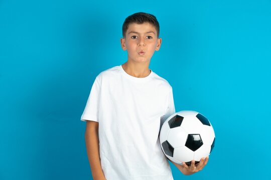 Shot of pleasant looking Little hispanic boy wearing white T-shirt holding a soccer ball , pouts lips, looks at camera, Human facial expressions