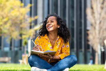 Happy young woman sitting on grass in park writing in book and looking away