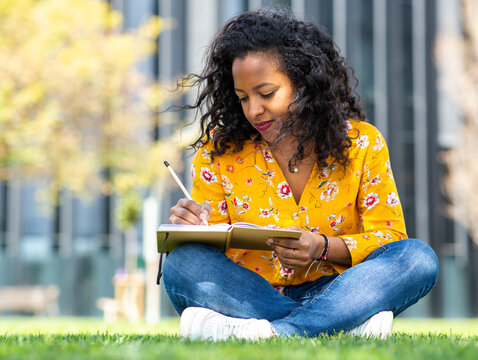Attractive young african woman sitting on grass outside and writing in book