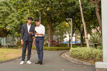 Obraz premium Two young asian men watch a basketball game or social media video on their cellphone while taking a walk at the park.