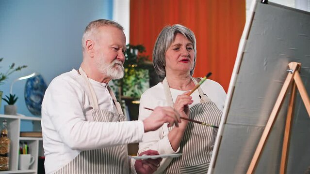 retired hobby, cheerful old man and woman in aprons with brushes and a palette in their hands paint a picture using an easel at home