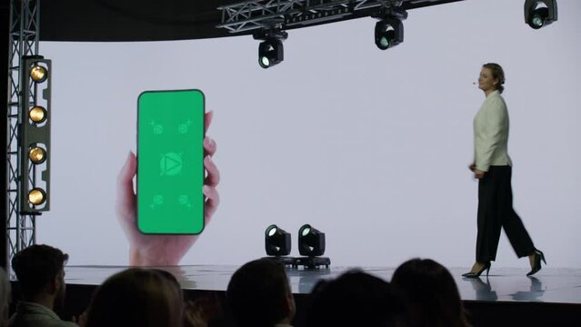 A woman presenter is entering a well-lit stage in front of a large LED screen showing a hand holding a generic smartphone with green screen. Technology, application or product launch presentation 