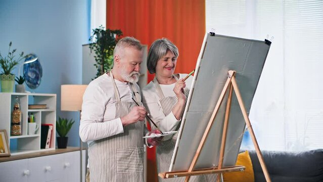 learning new skills in retirement, joyful old couple painting a picture with brushes and paints using an easel in the room