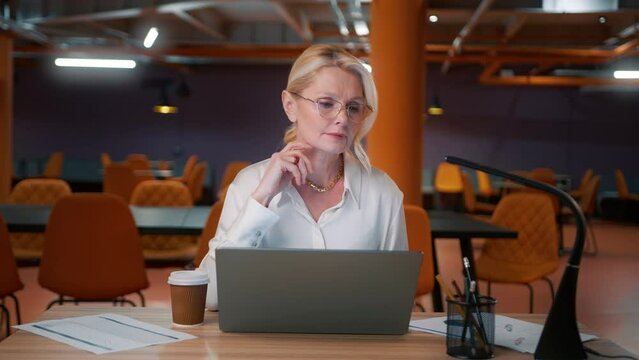 Mature attractive woman in eyeglasses working late. Lady boss working on project in modern loft style empty office on motion background. Business woman 50s typing an email on laptop, reading news 4K