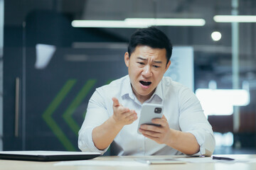 Angry young asian business man sitting at the desk in the office, holding the phone and looking worried and pointing at the screen.