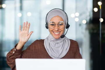 Close-up photo. Young hispanic female student in hijab sitting at desk in office wearing headset and talking on video call from laptop. He waves his hand and says hello to the camera on the monitor.