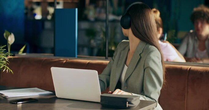 Calm attractive young woman in buiisness suit wearing headphones earbuds sits at table at cafe in front of laptop watches films cartoons interviews podcast during break from work job.