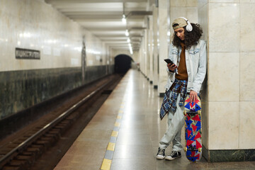 Teenage guy in headphones scrolling through playlist in smartphone while standing by railway,...