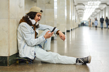 Side view of teenager with mobile phone texting, watching online video or looking through his playlist while sitting at subway station