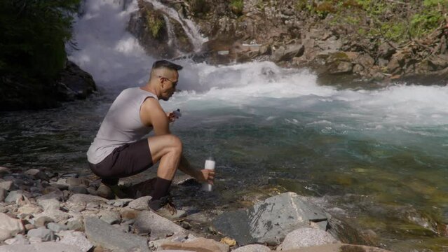 Man hiking and collecting spring water from river with his water bottle Wide Shot