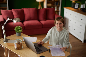 Smiling woman examining her bank statement at home office using laptop