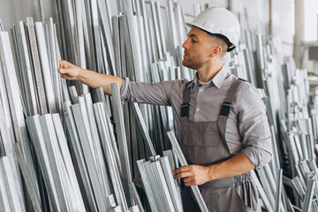 Happy bearded male factory worker in special uniform and white hard hat holding aluminum frame at production of metal plastic windows and doors