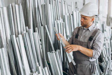 Happy bearded male factory worker in special uniform and white hard hat holding aluminum frame at...