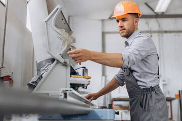 A male worker in a special uniform and an orange helmet works on a CNC machine for the production of PVC windows and doors at the factory.