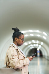 Profile view of young businesswoman in stylish trenchcoat using mobile phone while standing in front of camera against subway tunnel