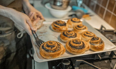 A female pastry chef removes freshly baked rolls sprinkled with poppy seeds and sugar from a baking sheet with a plastic spatula.