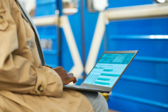 Laptop screen with graphic financial data beign analyzed by female broker in casualwear sitting at metro station against blue subway train