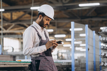 Happy bearded Indian male factory worker in special uniform and white hard hat holding the phone...